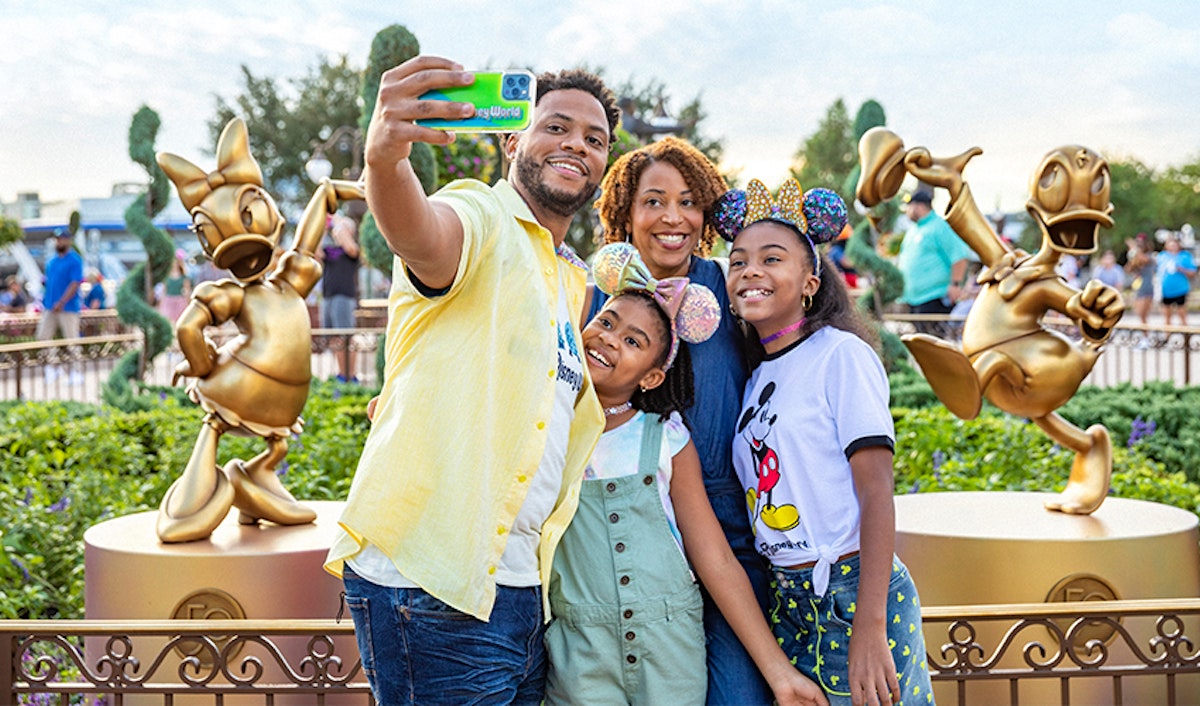 A family taking a selfie with a smartphone at an amusement park, with playful statues in the background.