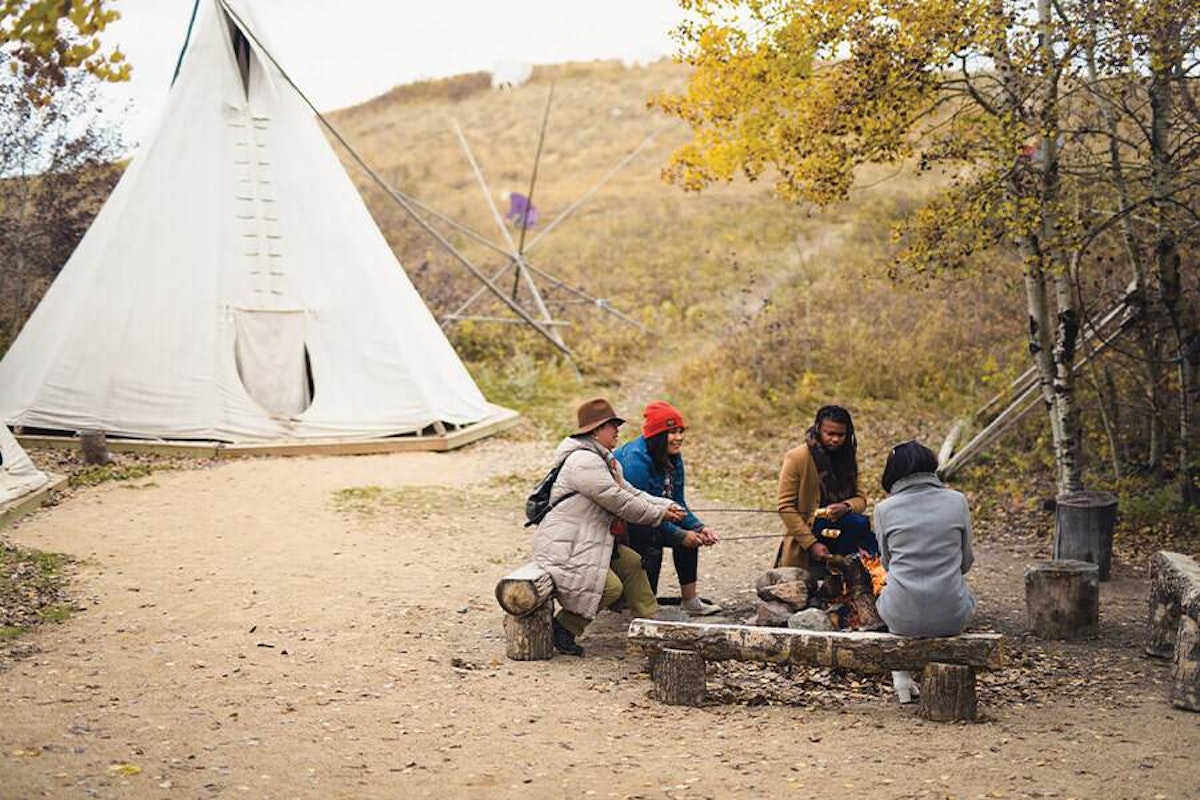 A group of people sitting around a campfire near a teepee.
