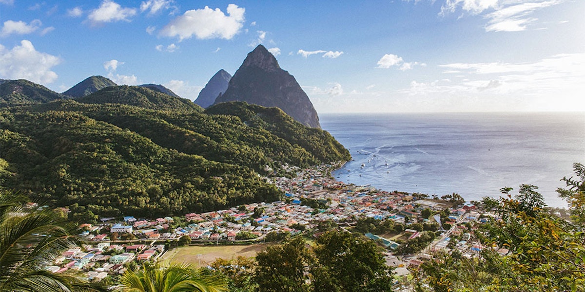 A coastal town nestled between lush green hills and the ocean, with towering peaks in the background.