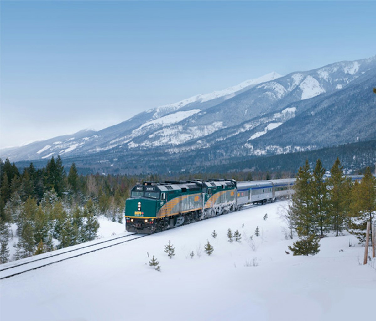 A passenger train travels through a snowy landscape with mountains in the background.