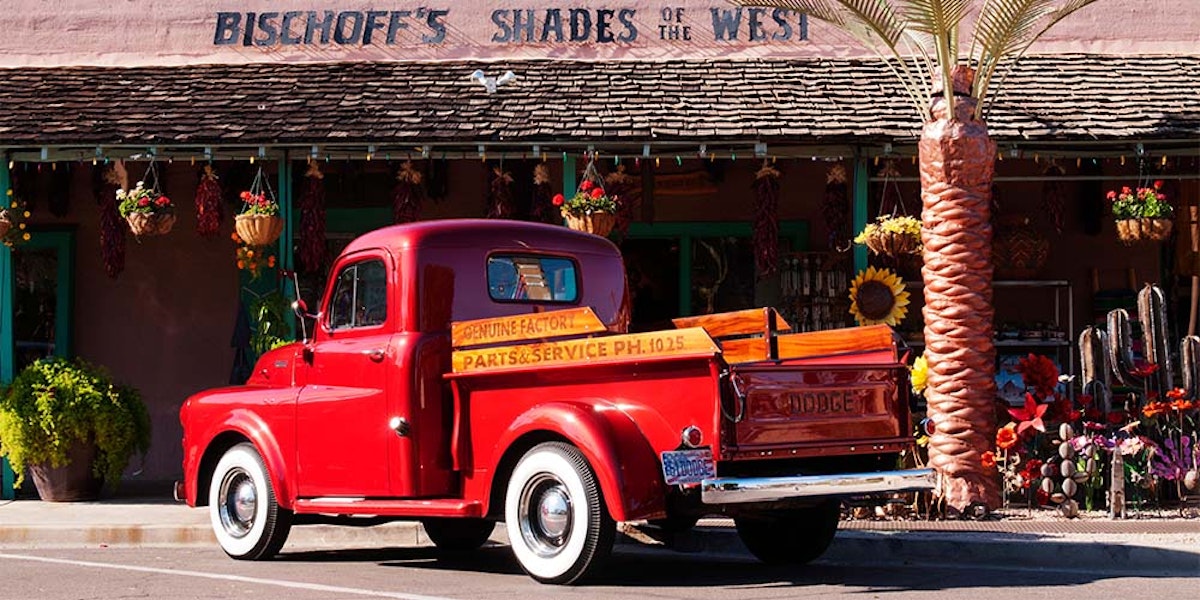 Vintage red pickup truck parked in front of a store with "bischoff's shades of the west" signage.