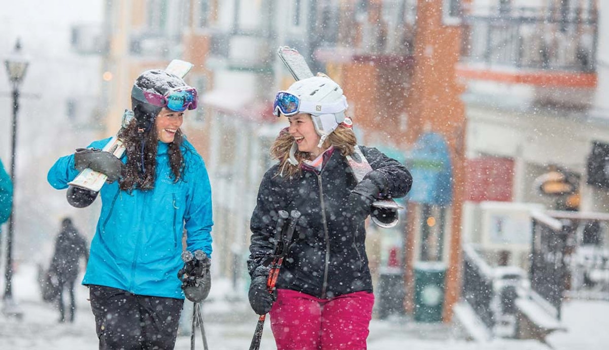 Two women in ski gear walking down a snowy street.