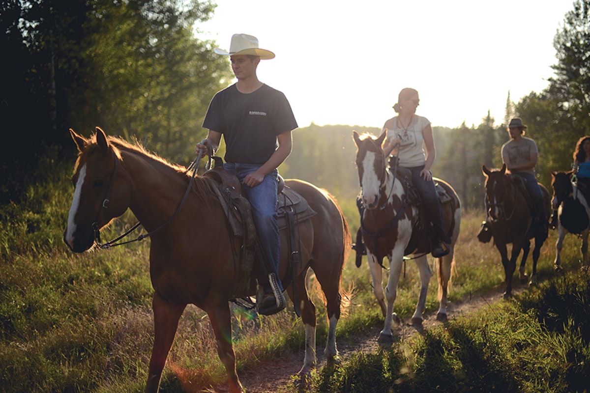 Group of people on horseback riding through a forest trail at sunset.