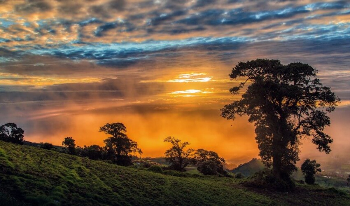 The sun is setting over a hill with trees and clouds.