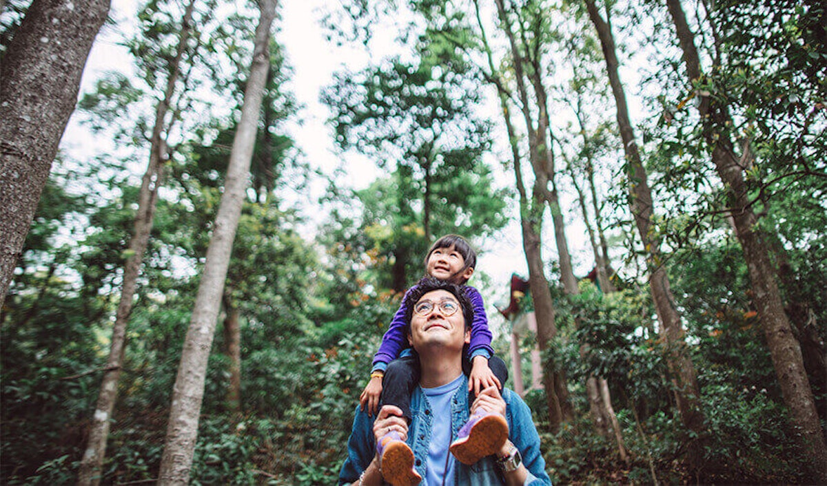 A child sitting on an adult's shoulders, both surrounded by trees.