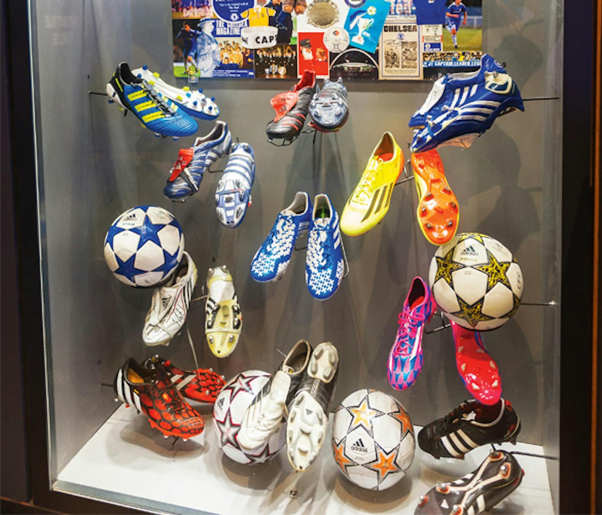 Display case of various soccer cleats and balls with memorabilia in the background.