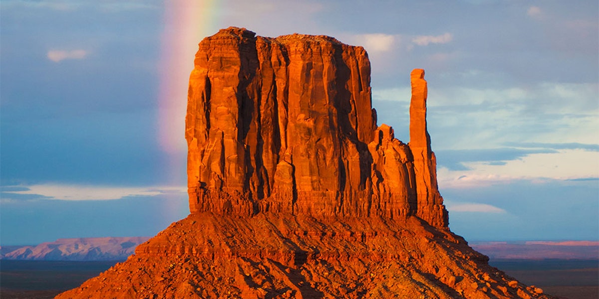 A rainbow appears beside a sunlit rock formation in monument valley at dusk.