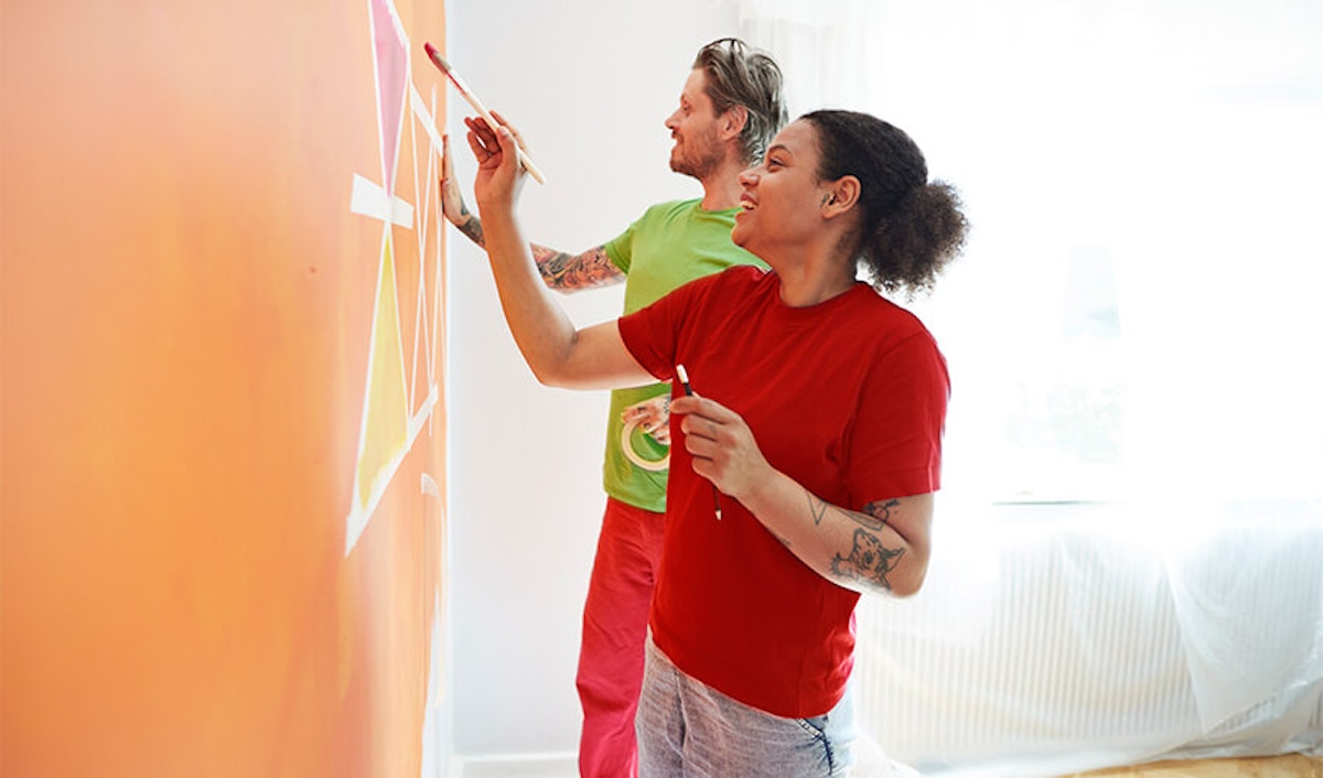 Two people painting an orange wall in a room.
