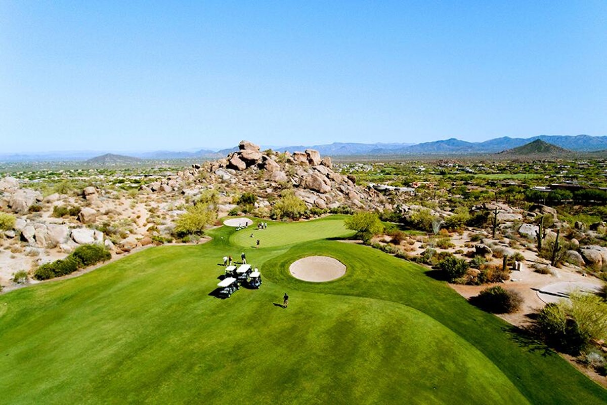 Aerial view of a desert golf course with golf carts and players on a green, surrounded by cacti and rugged terrain.