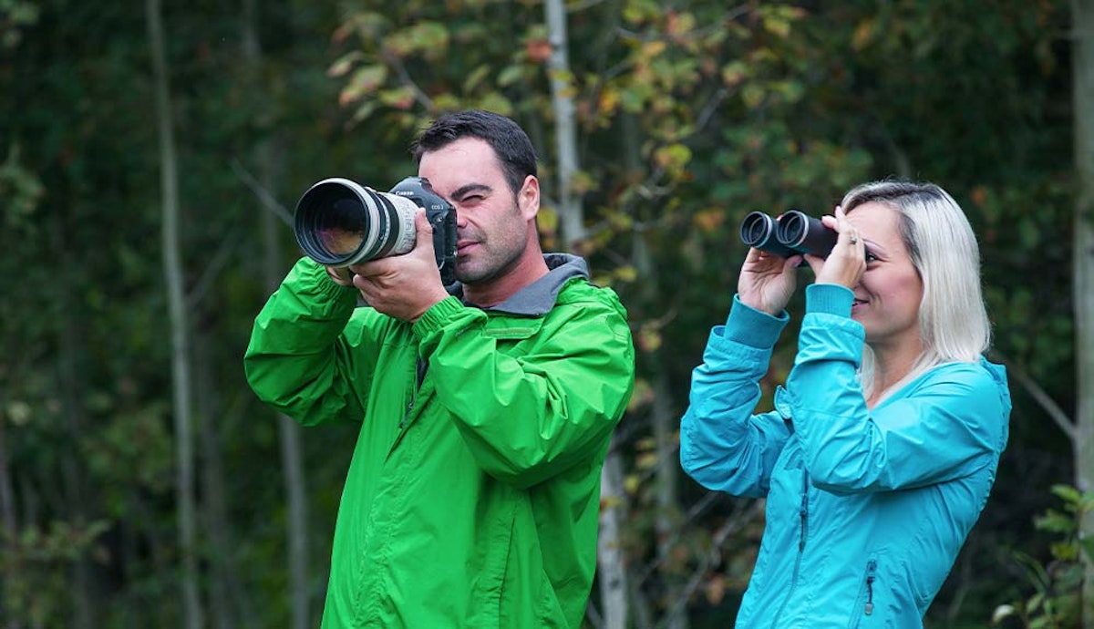 Two people looking through binoculars in the woods.