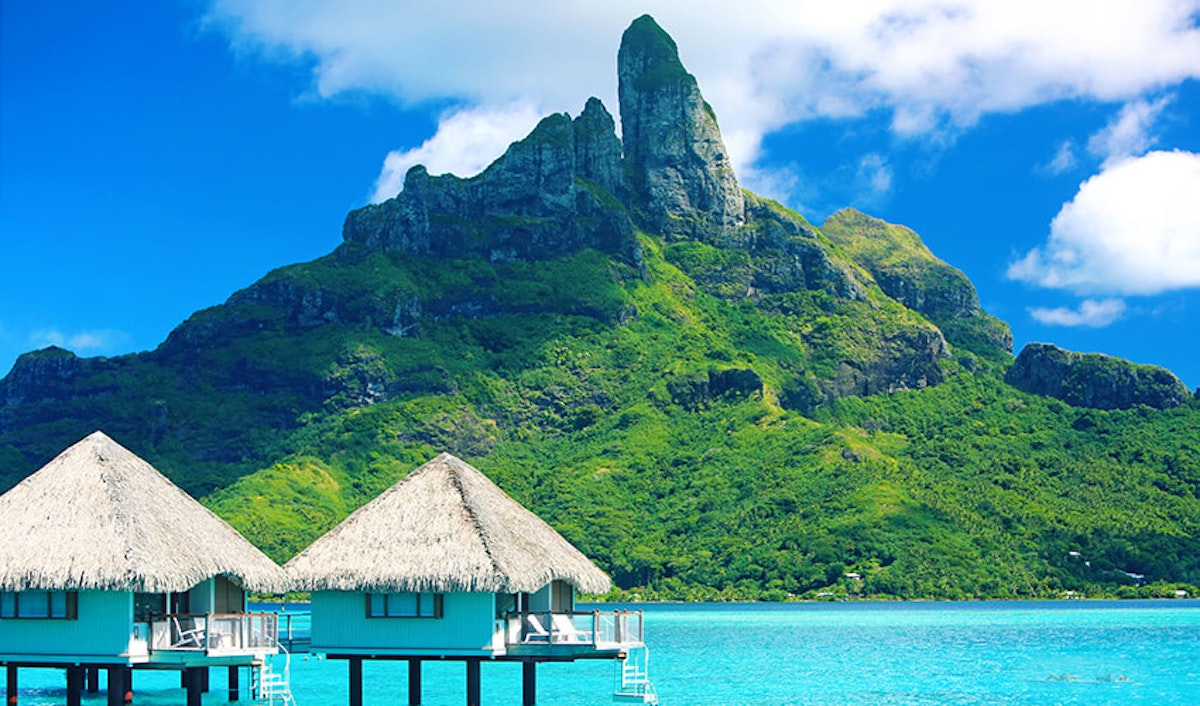 Overwater bungalows with thatched roofs facing a lush green mountain on a tropical island.