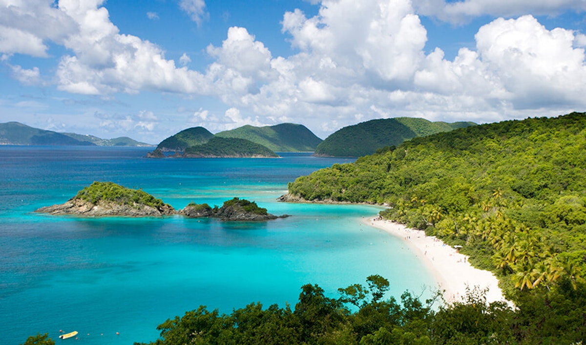 Tropical beach with clear turquoise waters, surrounded by lush green hills under a partly cloudy sky.