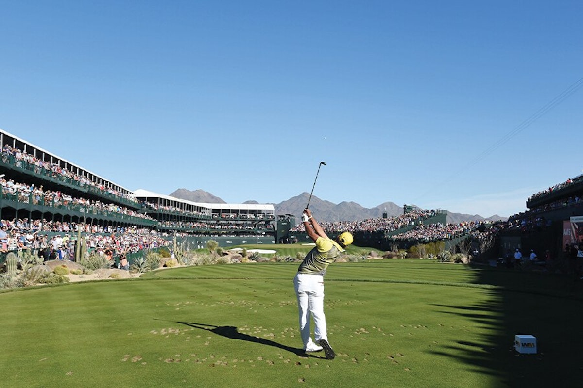 Golfer taking a swing on a course with spectators watching from stadium seating against a backdrop of mountains.