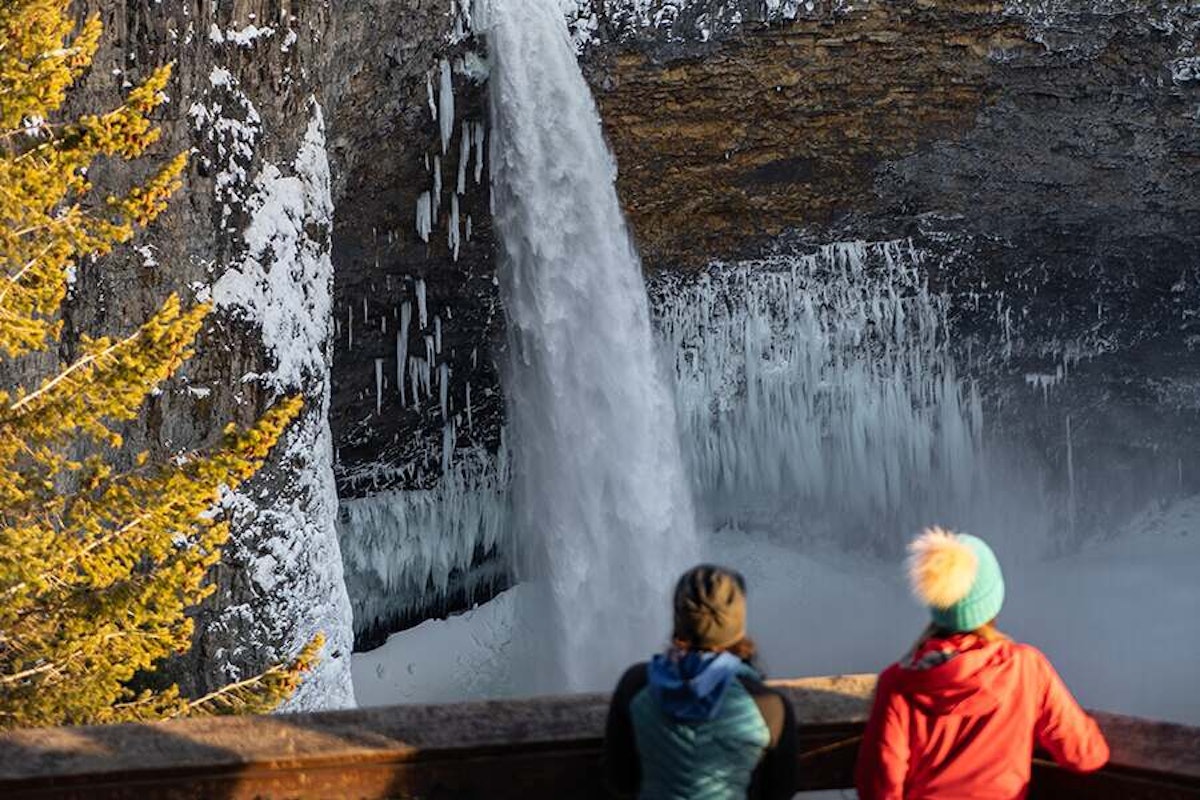 Two people observing a partially frozen waterfall in a wintry landscape.