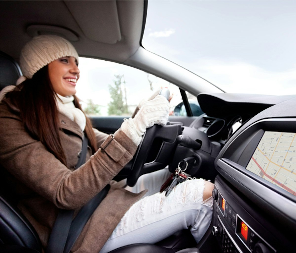 A smiling person wearing a winter hat and scarf driving a car with a navigation system on the dashboard.