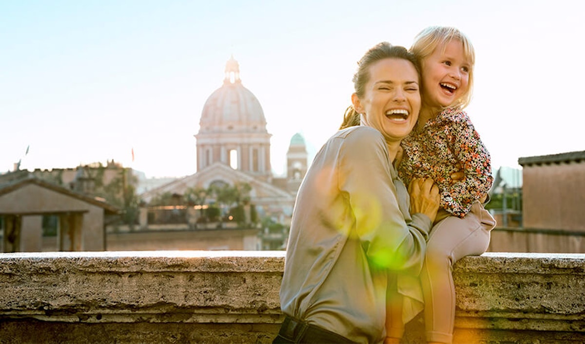 A woman and a child smiling joyfully with a cityscape and a cathedral dome in the background during sunset.