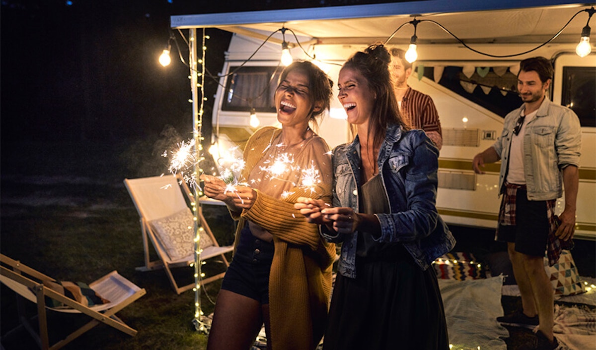 Two women holding sparklers and laughing at a night-time outdoor gathering with a camper van in the background.