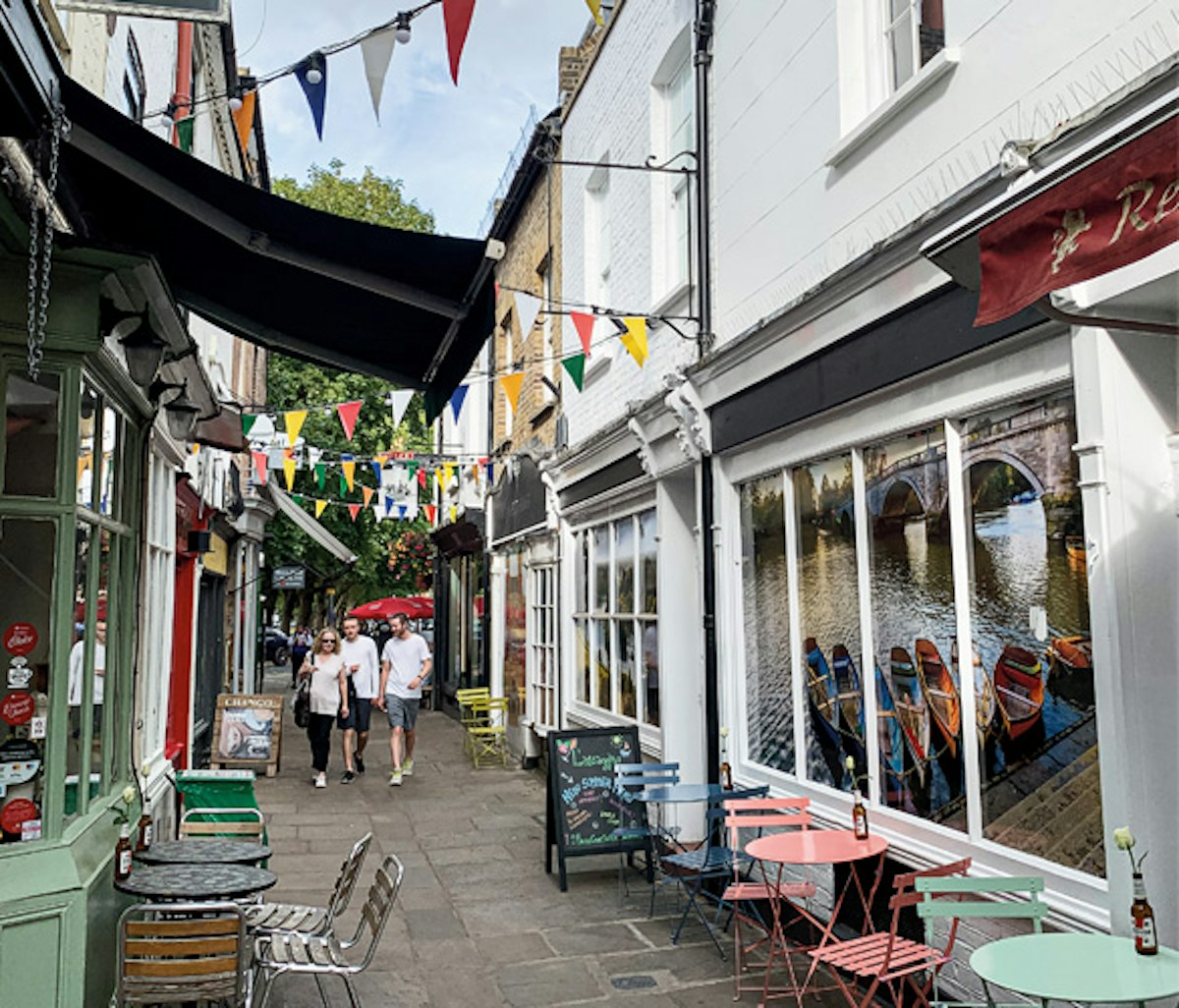 Quaint street with colorful bunting, outdoor seating, and canal boats visible in the background.