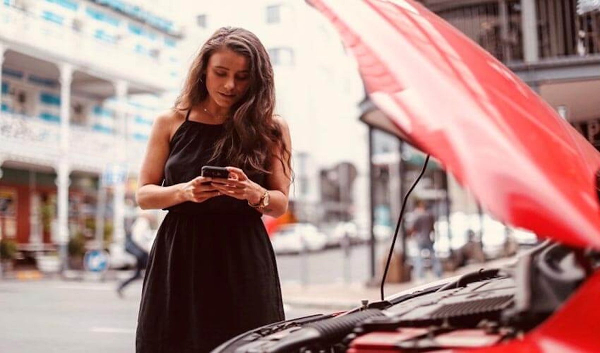 A woman looking at her phone while standing next to a red car with its hood open.