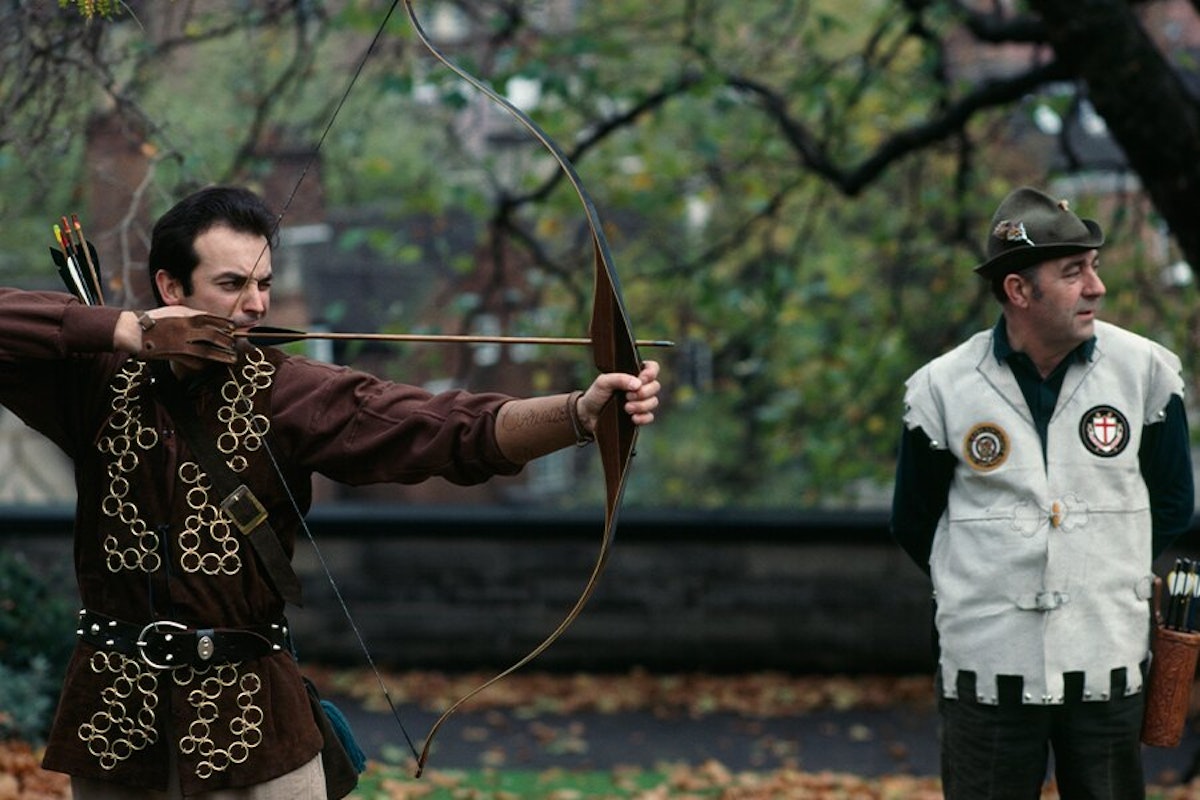 A person dressed as an archer aiming a bow and arrow, with a spectator in a ranger-like uniform watching closely.