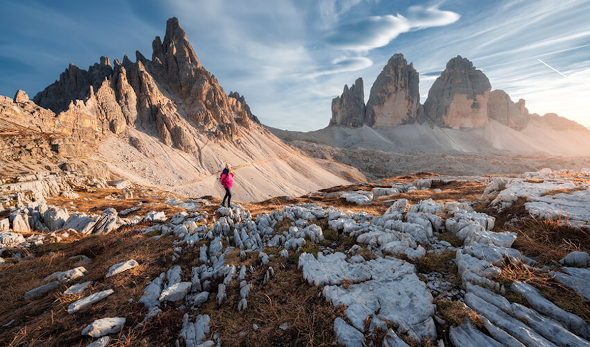 A hiker in a pink jacket walking towards the jagged peaks of the tre cime di lavaredo under a clear sky.