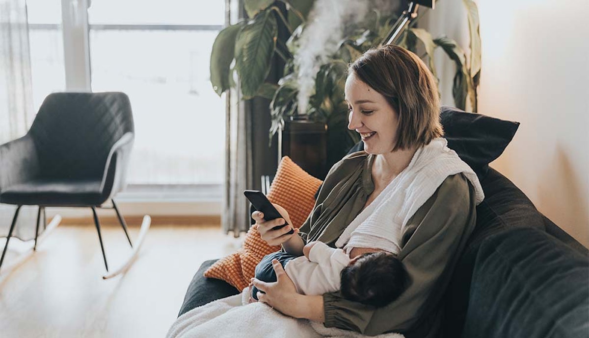 A woman is sitting on a couch and holding a baby while using a cell phone.