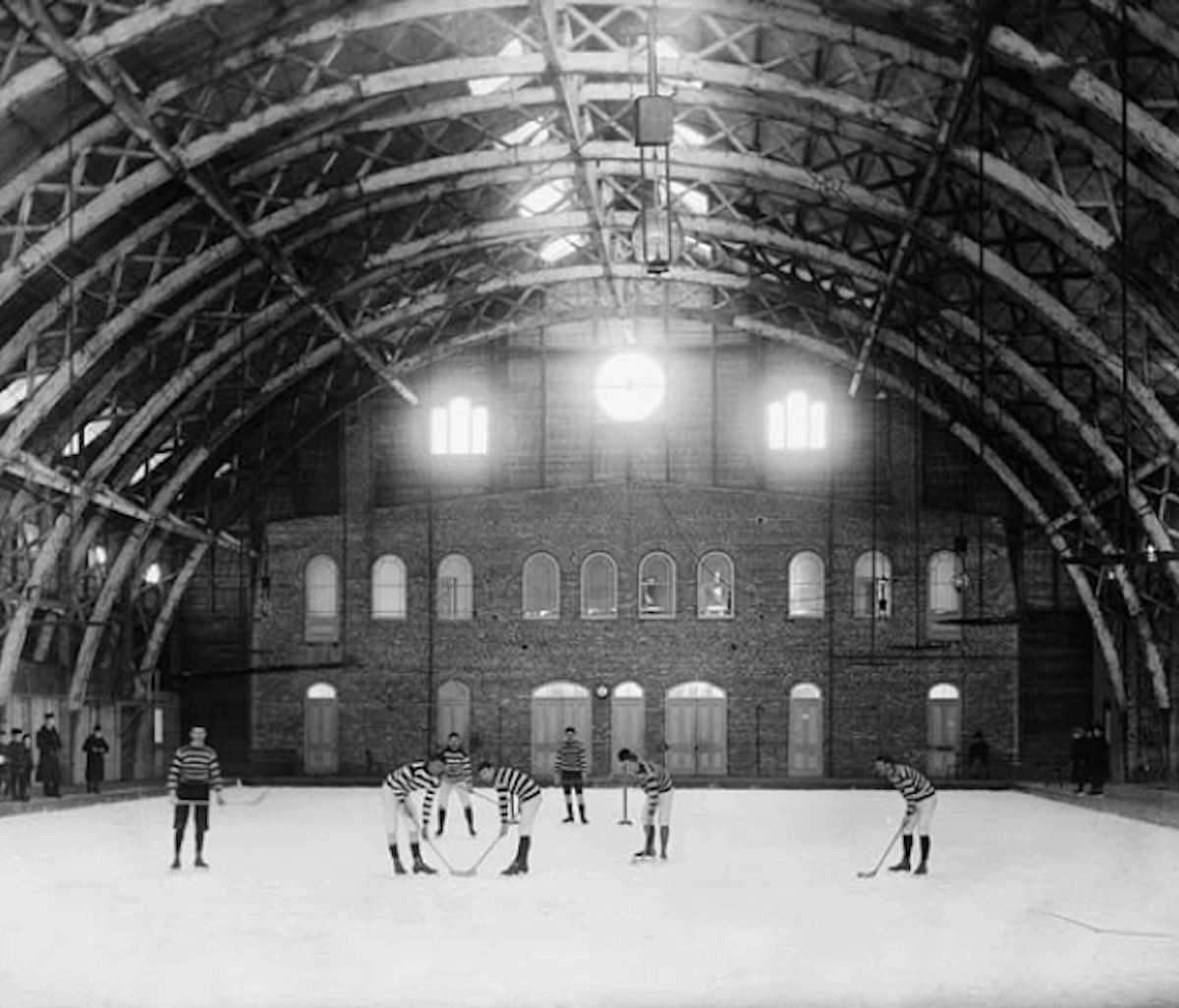 Referees on an ice rink inside a vintage arched-roof building.