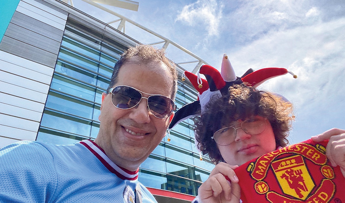 Two soccer fans posing in the sunshine, with one wearing a jester hat and holding a manchester united scarf.
