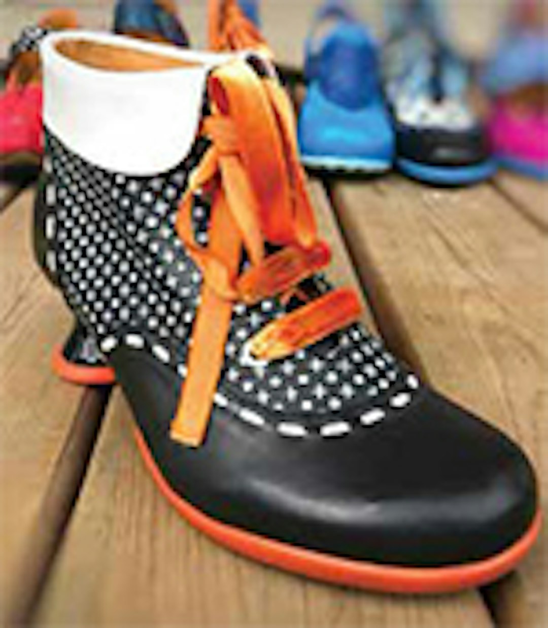 A stylish black and white patterned shoe with orange laces, displayed on a wooden floor with a blurred background of various other shoes.