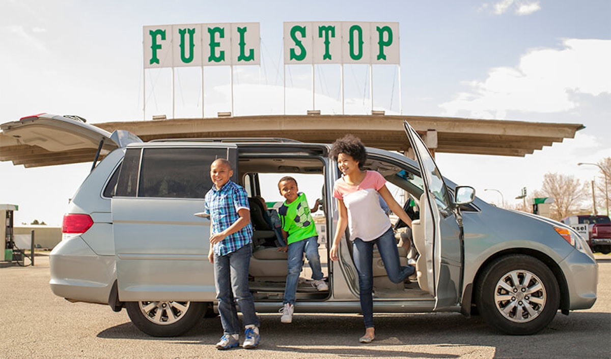 A family getting out of their minivan at a fuel stop on a sunny day.