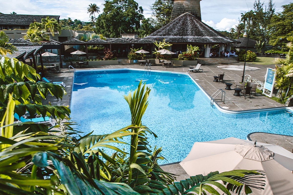 A tropical resort pool surrounded by lush greenery and lounge areas under a clear sky.