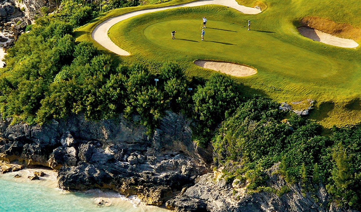 Aerial view of golfers on a scenic coastal golf course with undulating green and adjacent rocky shoreline.