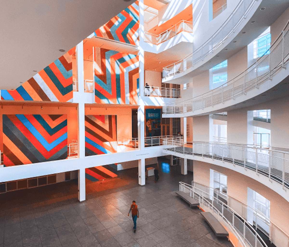 Modern building interior with a colorful striped wall art installation and a lone individual walking through the spacious atrium.