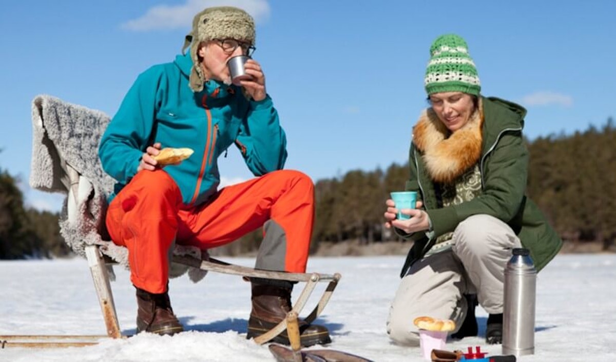 Two people sitting on a chair in the snow with a cup of coffee.
