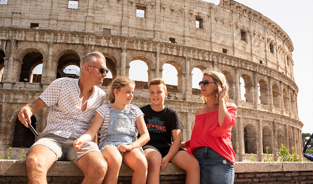A family of four sitting and smiling in front of the colosseum in rome.