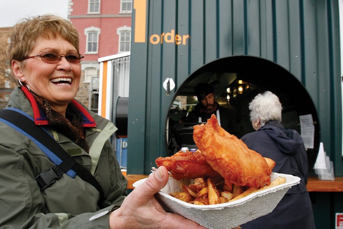 Woman smiling with fish and chips in front of a food stall.