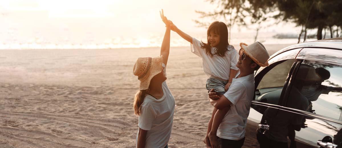 A mother, father and young daughter stand beside a car at the beach as the sun sets behind them.