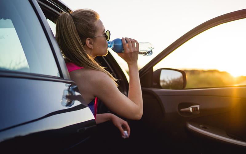 Woman with an open car door drinking water and looking at a setting sun