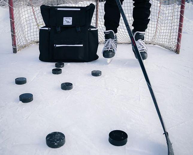 The lower half of the legs of someone wearing skates standing on ice with a hockey net behind them and a black hockey bag beside them. They hold a hockey stick in front of them and there are hockey pucks scattered on the ice.
