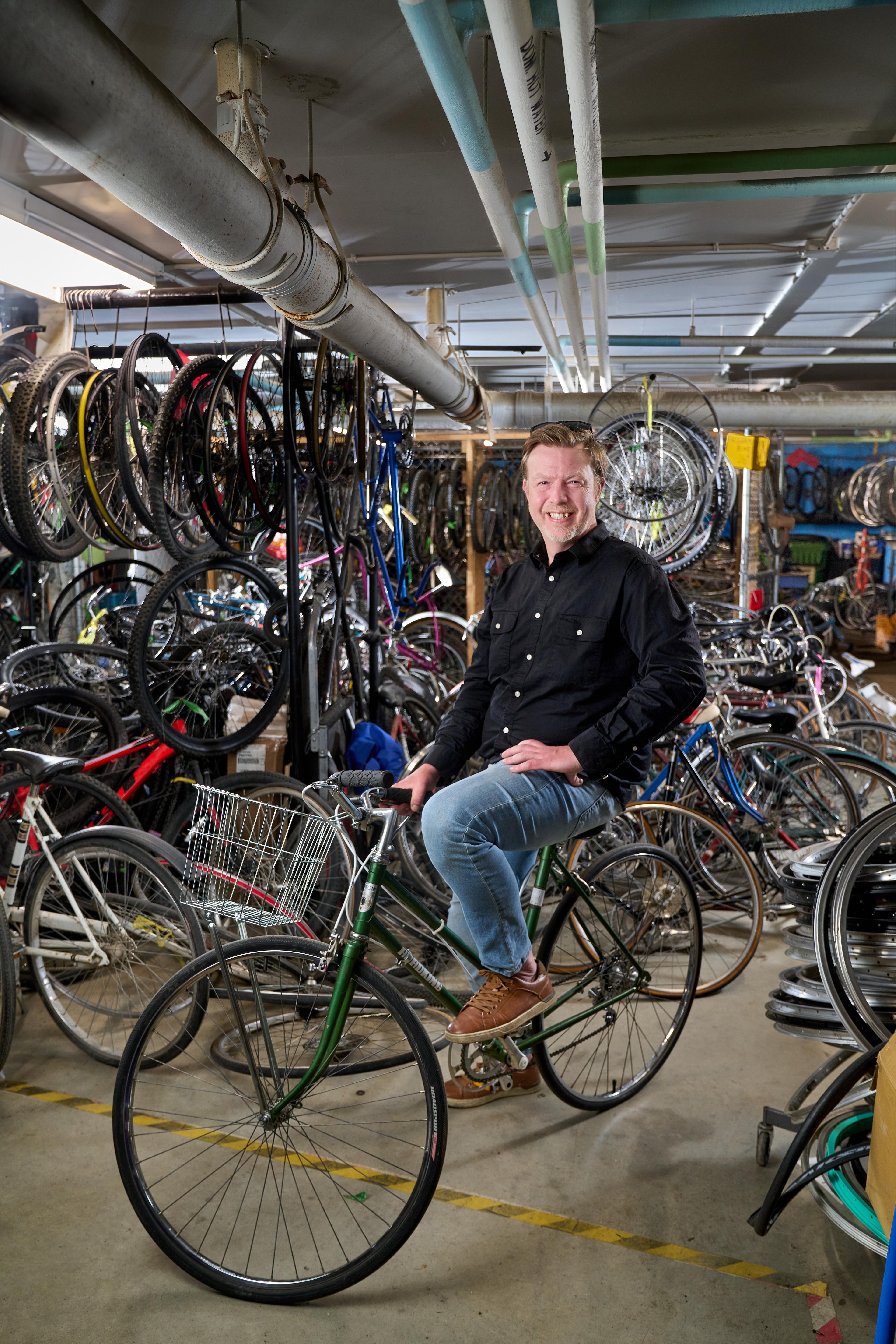 A man wearing blue jeans and a black button-up shirt smiles and sits on a bike in the middle of a warehouse with dozens of bikes behind him.
