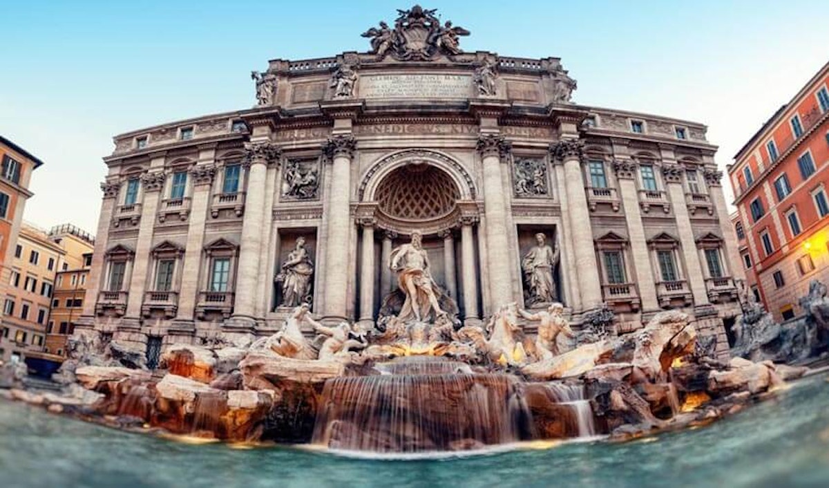 A large stone building with a fountain and statues with trevi fountain in the background.