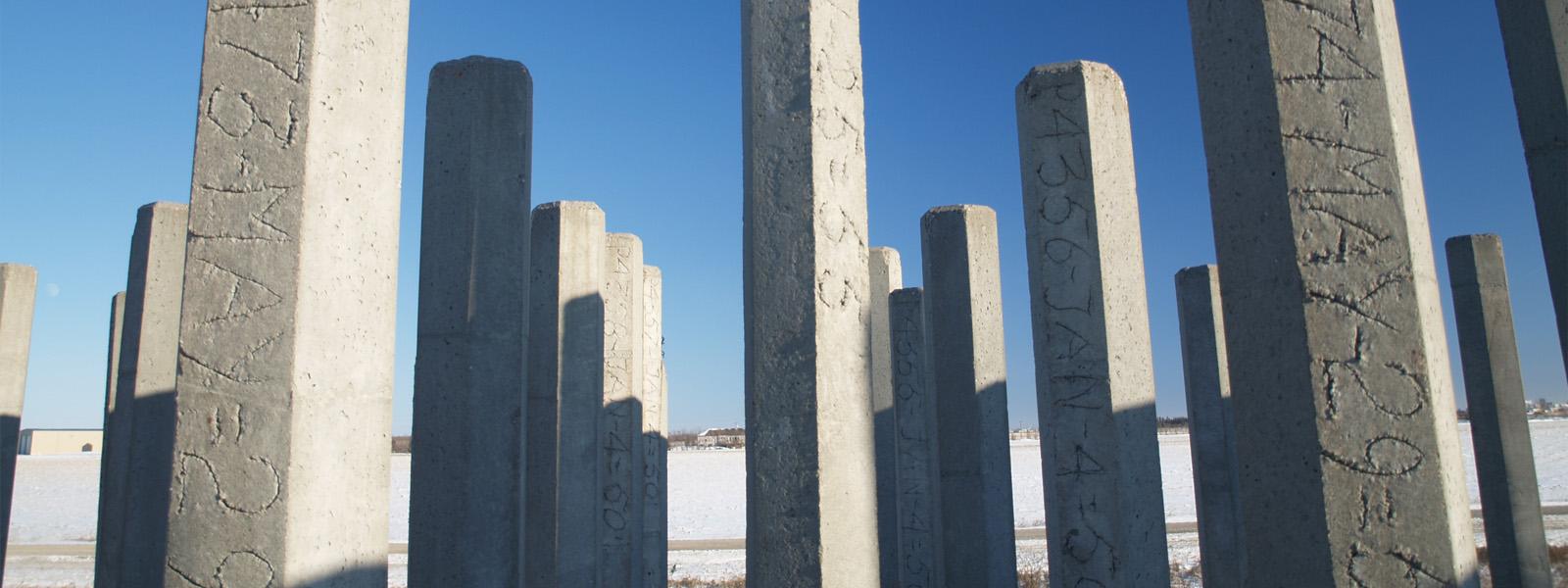 Dozens of grey, cement poles sticking up out of the snowy ground. The sky above them is clear and there are random letters and numbers inscribed on each pole.