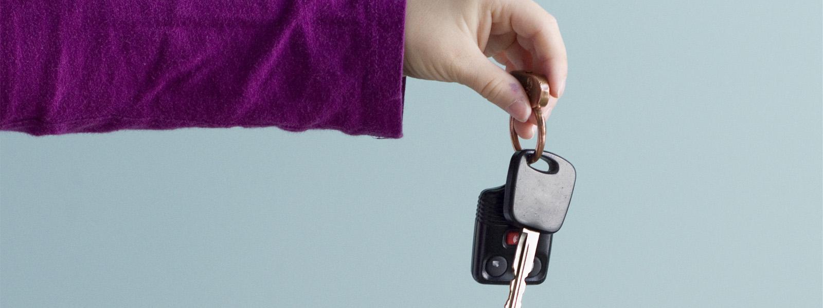 A person off-camera outstretches their arm, wearing a purple long-sleeved shirt, and holds a keyring with a car key on it.