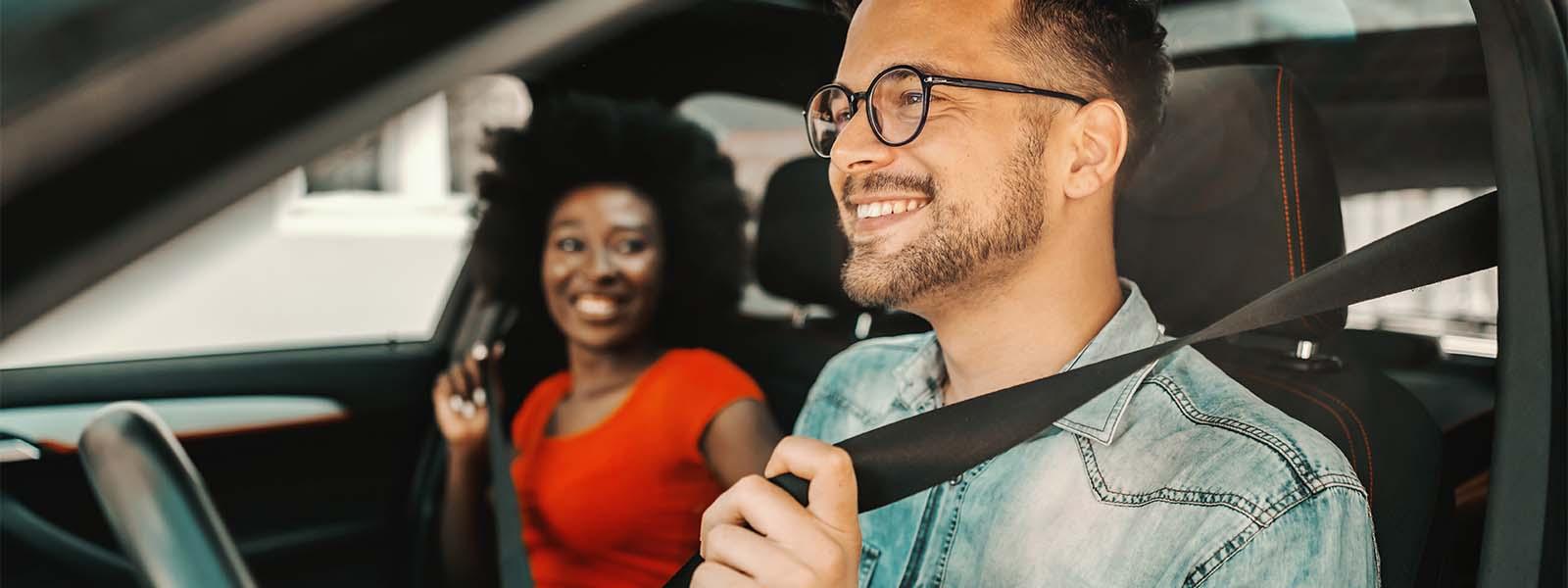 A white man in a denim shirt wearing glasses with facial hair is smelling while in the driver’s seat of a vehicle. In the passenger seat next to him is a Black woman looking at him smiling, wearing a red T-shirt and has an Afro.