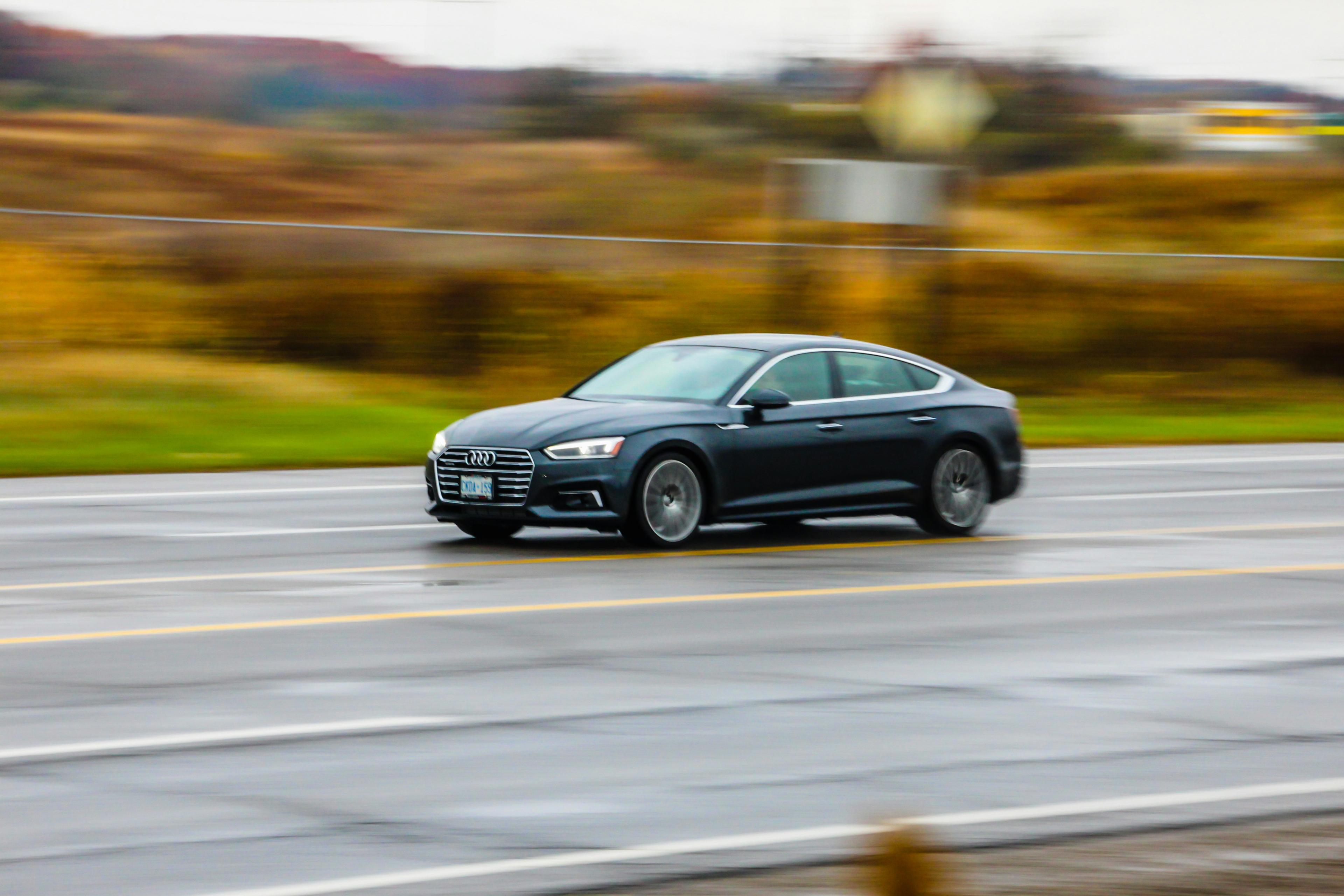 A black car driving down a road. It is implied that the car is driving fast because its surroundings are blurry.