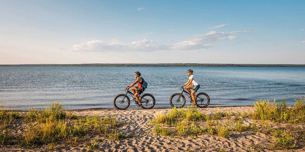 Two people riding bikes on the beach near a body of water.