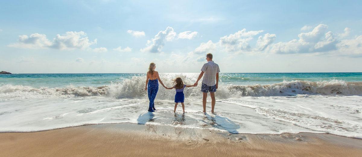 A family of three, a mother, father and young daughter, standing in at the beach holding hands. They are standing at the edge of the water as the waves are approaching them.