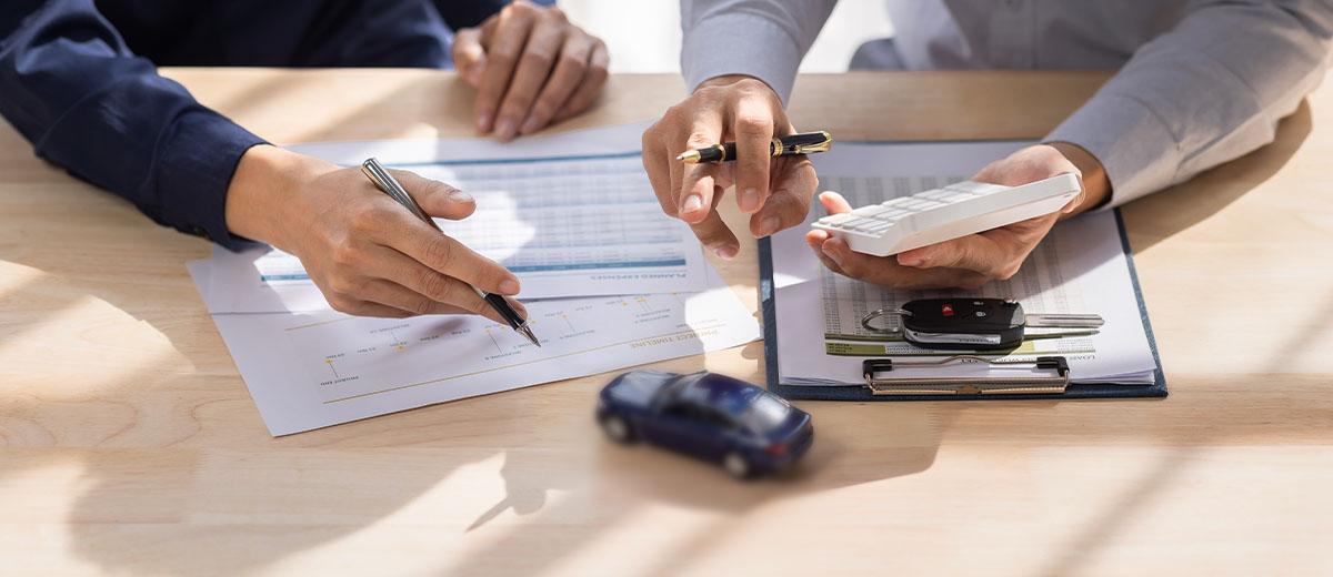 A close-up shot of the hands of two people sitting at a desk. One person is holding a calculator while the other is holding a pen. Below their hands are various sheets of paper.