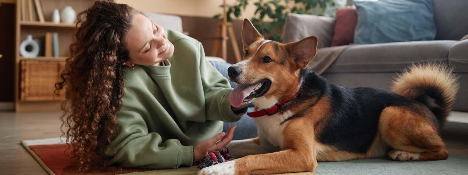 A woman with long light brown curly hair is lying on her side on the floor of her living room next to her dog, who looks happy. The dog’s tongue is hanging out of its mouth.
