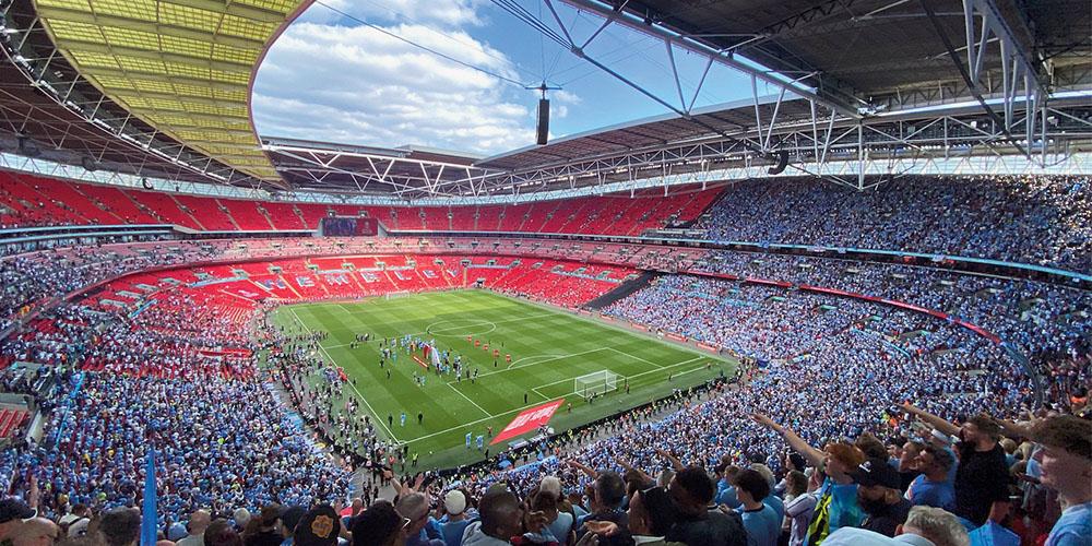 A crowded stadium with fans in blue and red during a sunny day, likely before a football match.